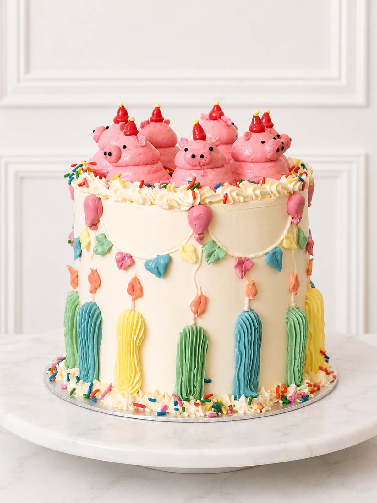 Full view of a playful birthday cake decorated with hand-piped pink pigs, party hats, pastel bunting, balloons, candles and sprinkles.