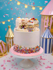 The Meta Funfetti Pinata Cake features white buttercream, colorful sprinkles at the base, and assorted cupcakes and sweets on top. It’s set against a pastel circus-themed backdrop with string lights, confetti, and a blue wall.