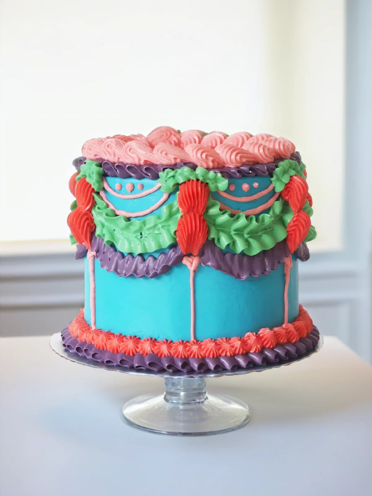 The Bright Lambeth Vintage Cake features pink, purple, green, and orange buttercream piping on a turquoise base atop a glass cake stand, bringing vibrant Bridgerton-style charm to any setting.