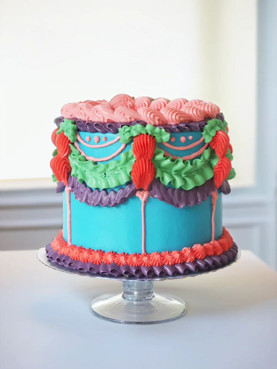The Bright Lambeth Vintage Cake features pink, purple, green, and orange buttercream piping on a turquoise base atop a glass cake stand, bringing vibrant Bridgerton-style charm to any setting.