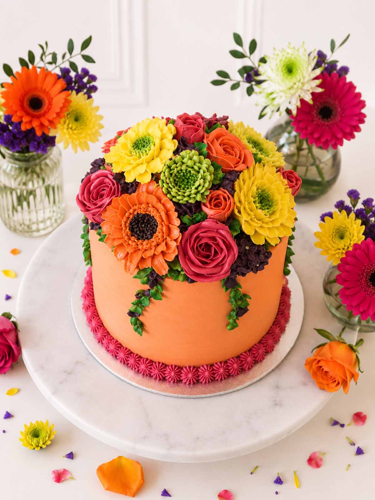 Bright floral buttercream cake surrounded by flowers on a marble surface