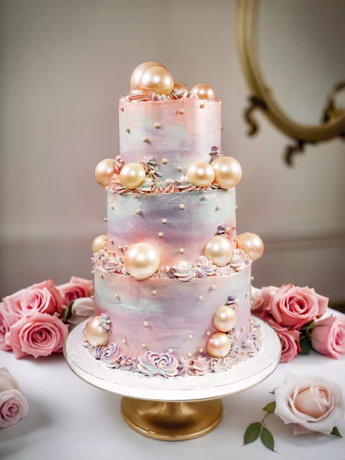 Three-tier cake with pastel watercolor frosting, gold pearls, and small white sprinkles, surrounded by pink roses