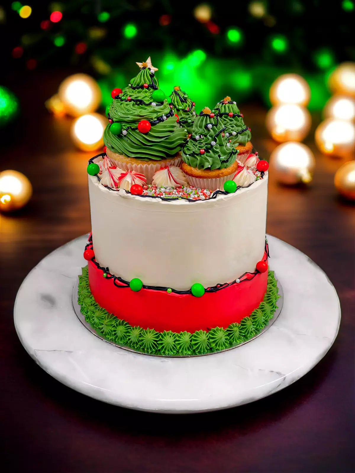 The Christmas Tree Cake features a vanilla sponge base, topped with cupcakes shaped like Christmas trees and decorated with red and green icing, served on a marble plate with blurred holiday lights in the background.