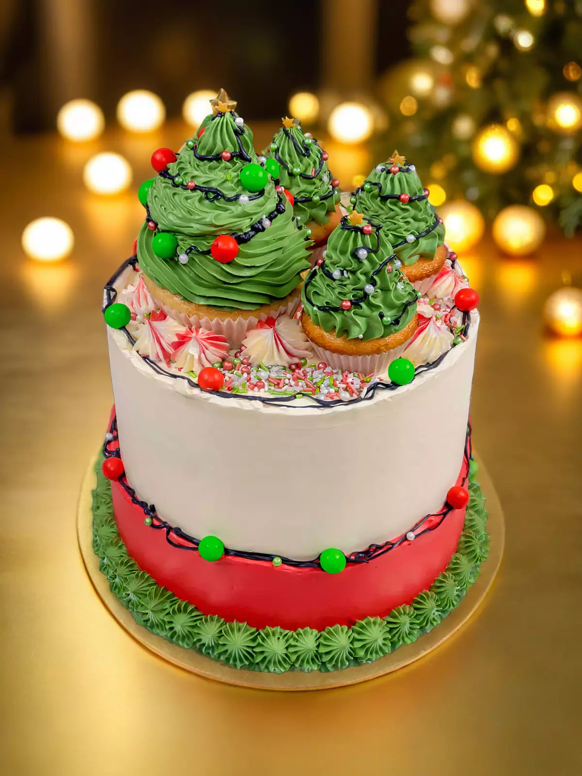 The Christmas Tree Cake features vanilla sponge topped with cupcakes decorated as Christmas trees, green frosting, red and green ornaments, and a red-and-white base, set against shimmering blurred lights and a glowing Christmas tree.