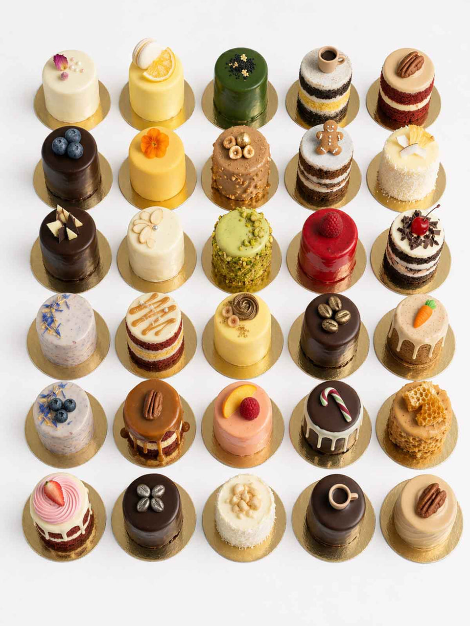 An assortment of 30 mini cakes, each with unique toppings and designs, arranged in rows on a white surface