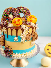 The Emoji Cake features blue and yellow vanilla Swiss meringue buttercream, topped with donuts, cookies, chocolate drizzle, smiling emoji macarons, and white frosting flowers on a gold cake stand.