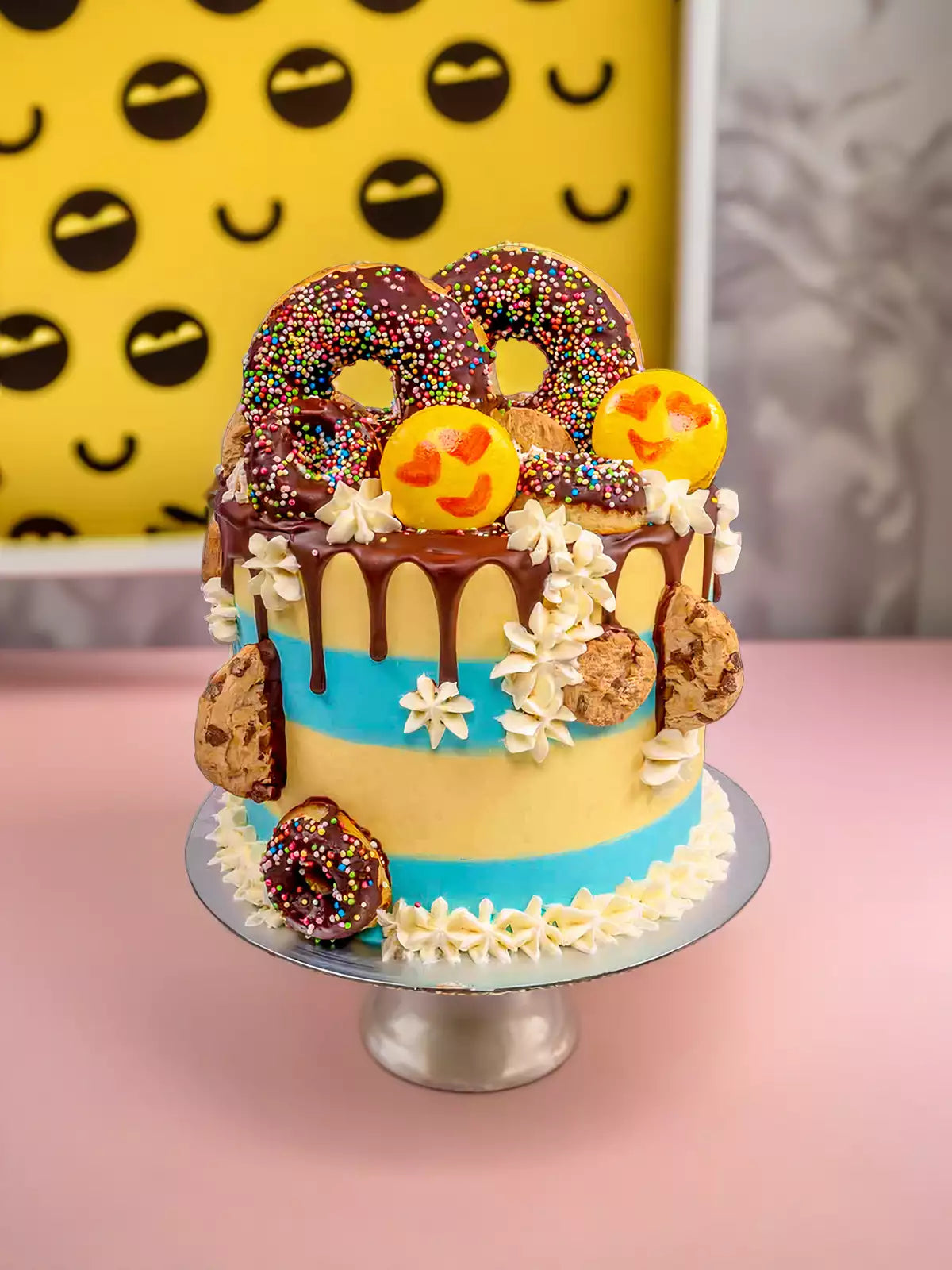 The Emoji Cake features chocolate-drizzled donuts, emoji macarons, chocolate chip cookies, and sprinkles on white vanilla Swiss meringue buttercream with blue icing stripes, showcased against a yellow emoji pattern background.