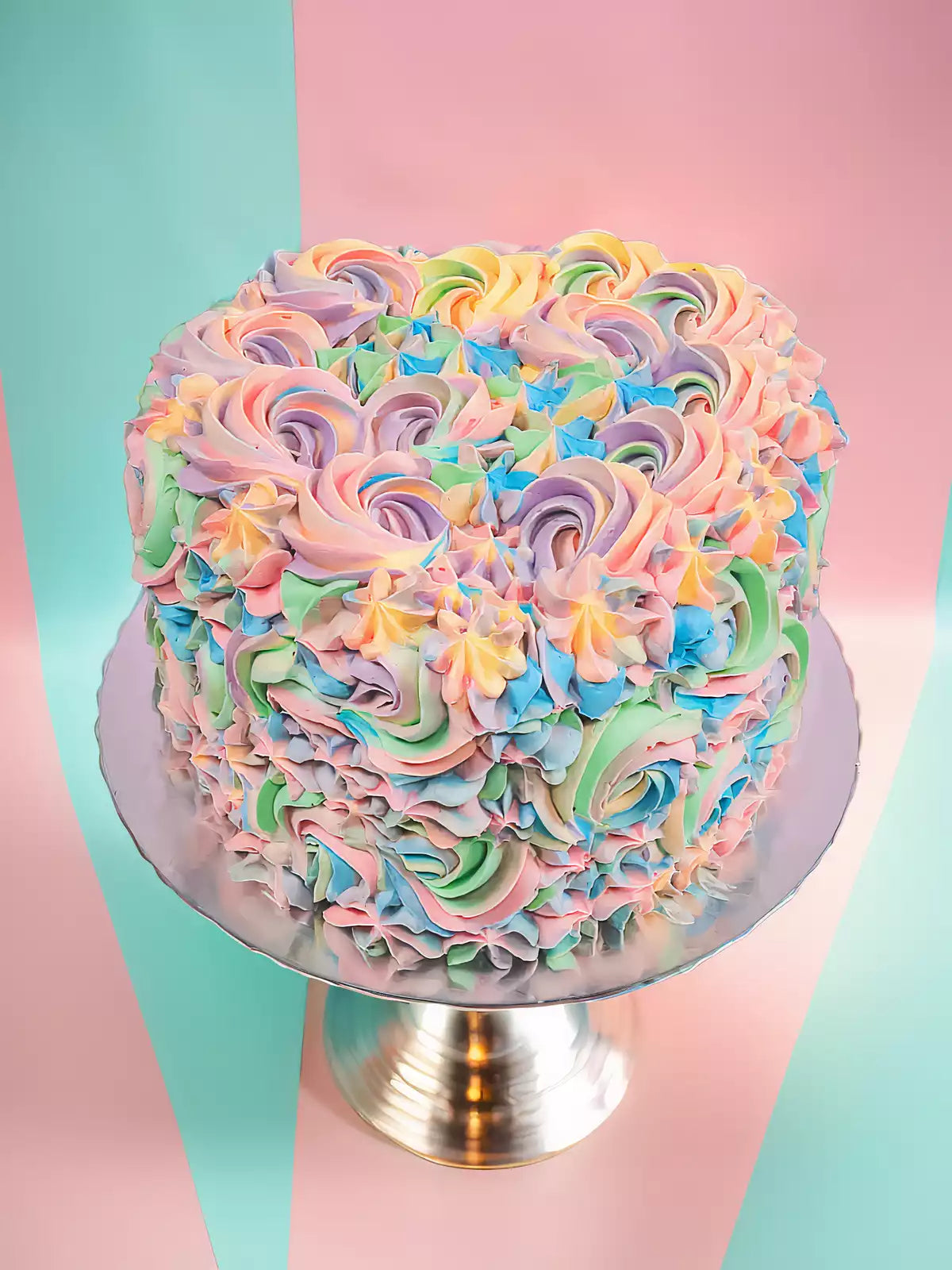 The Pastel Swirl Heart Cake features pastel rainbow Swiss meringue buttercream rosettes and sits on a silver cake stand against a mint green and pink background.