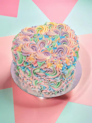 The Pastel Swirl Heart Cake is a round vanilla sponge topped with pink, purple, blue, and green Swiss meringue buttercream swirls on a silver board against a vibrant geometric background.