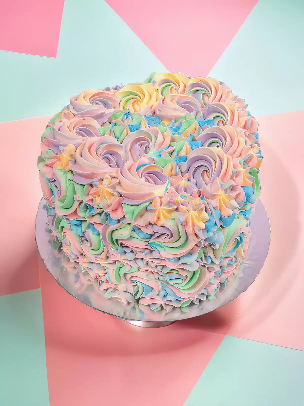 The Pastel Swirl Heart Cake is a round vanilla sponge topped with pink, purple, blue, and green Swiss meringue buttercream swirls on a silver board against a vibrant geometric background.