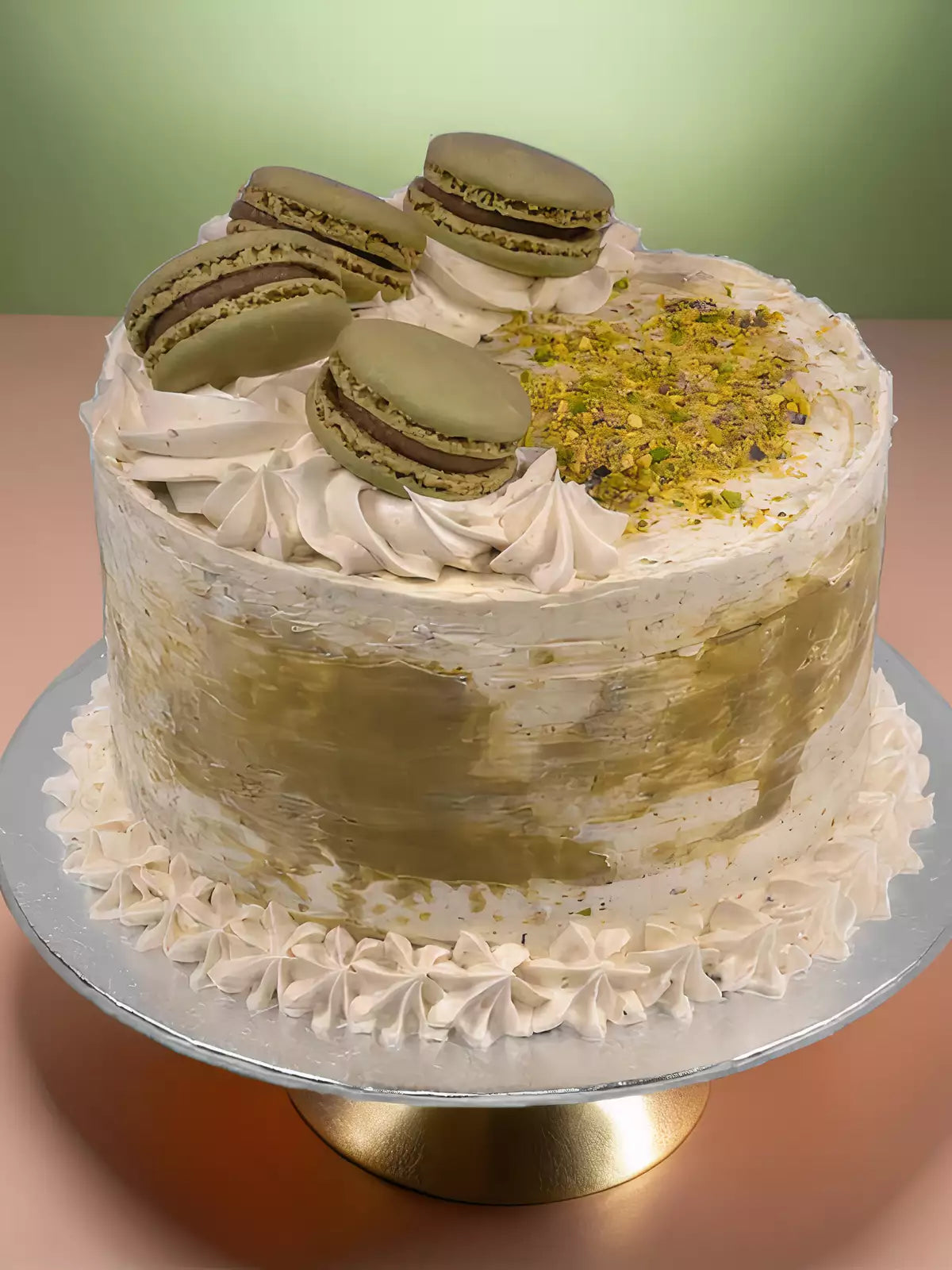 The Orange and Pistachio Cake is a round Mediterranean dessert with white and greenish frosting, topped with four green macarons and crushed pistachios. It sits on a silver platter with piped cream around the base.