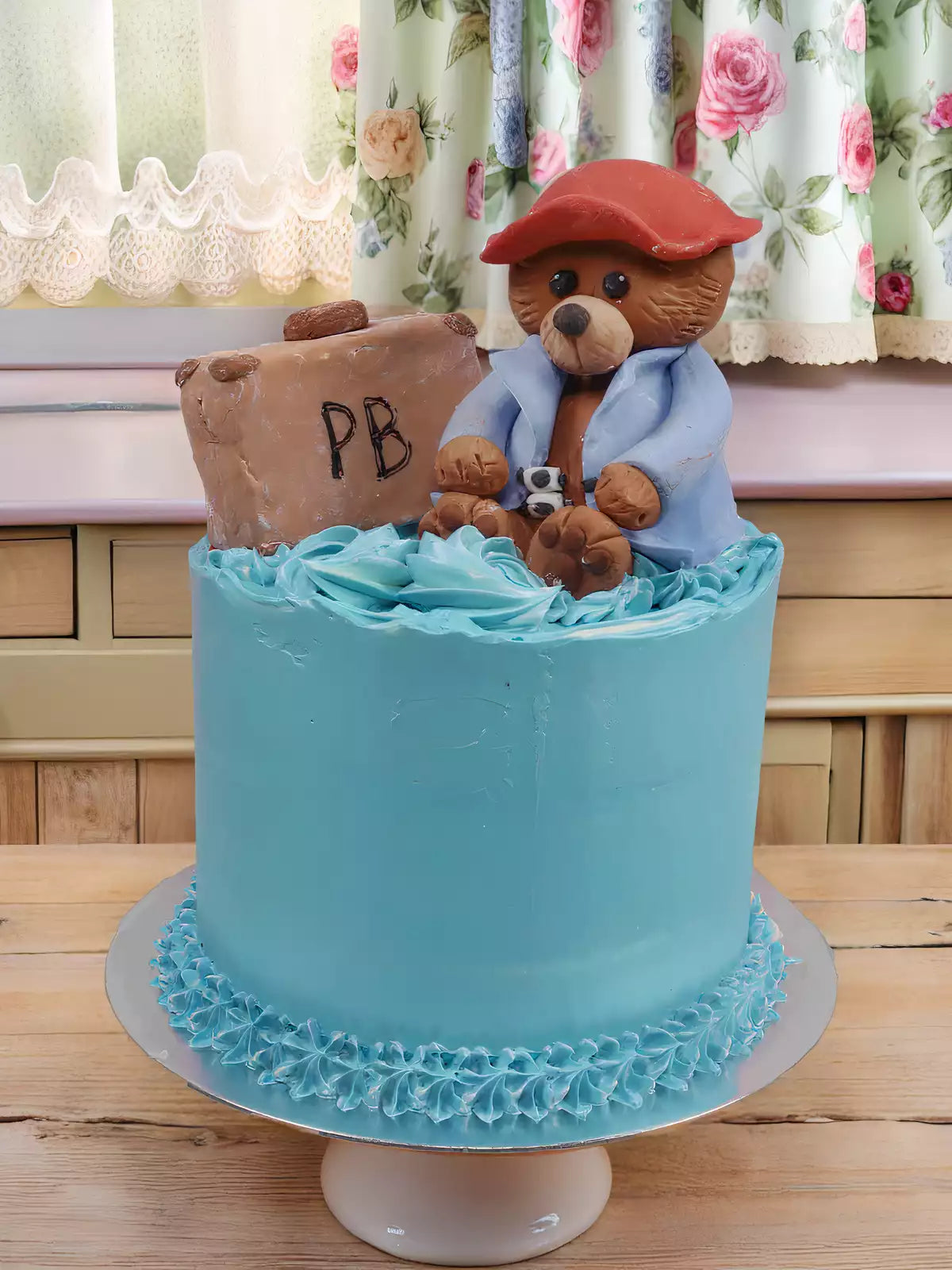 Bear Cake: A blue vanilla sponge cake featuring a handmade sugar paste bear in a red hat and blue coat holding a sandwich, with a brown "PB" suitcase beside him. Displayed on a white stand with floral curtains in the background.
