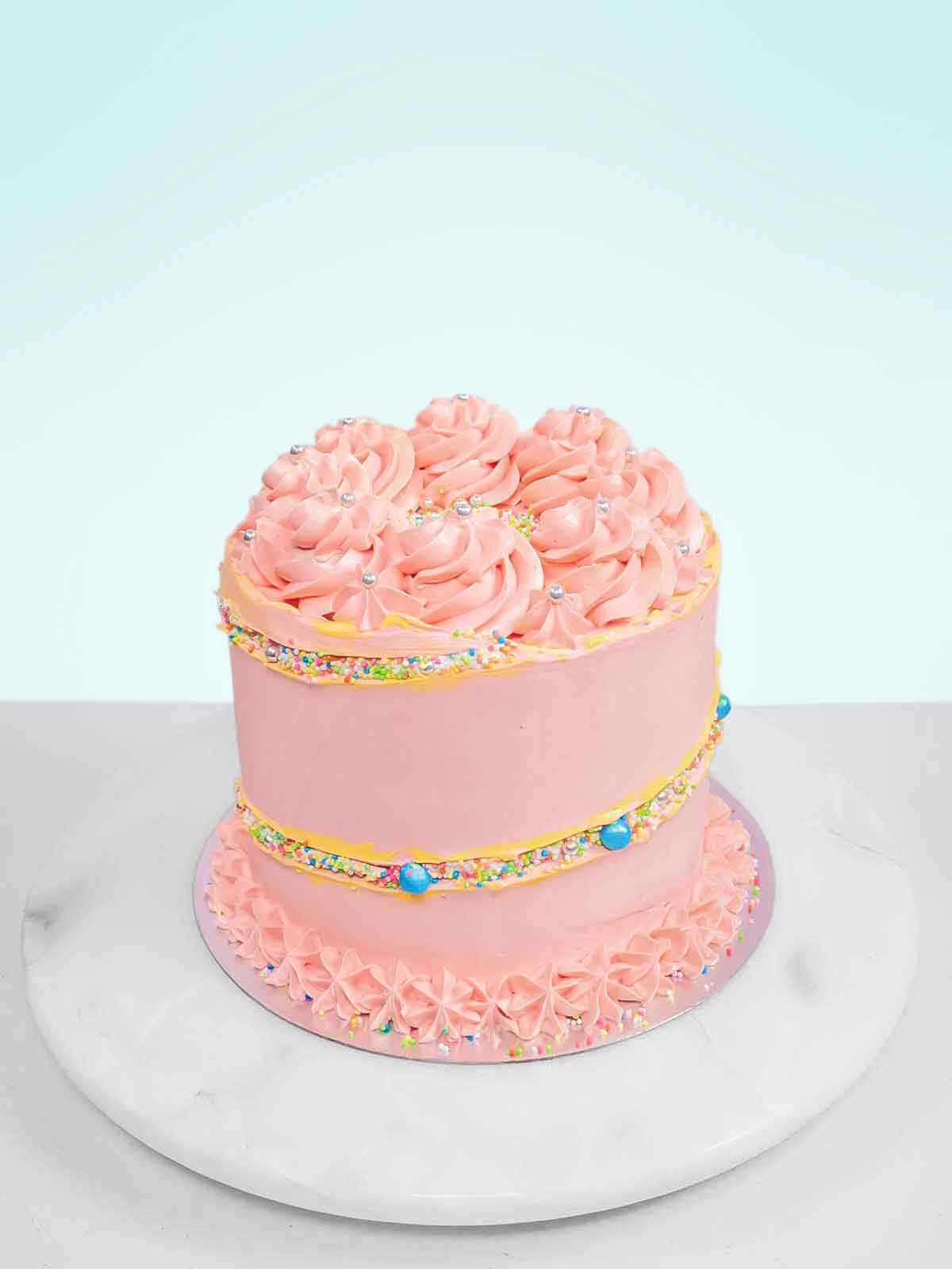 Pink cake with frosting rosettes, silver beads, sprinkles, and blue candies on a white marble stand