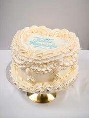 The Custom Registry Wedding Cake is heart-shaped with ornate frosting borders, seashell decorations, and light blue icing. Displayed on a gold cake stand, this personalized "Just Married" cake adds a charming touch to your celebration.