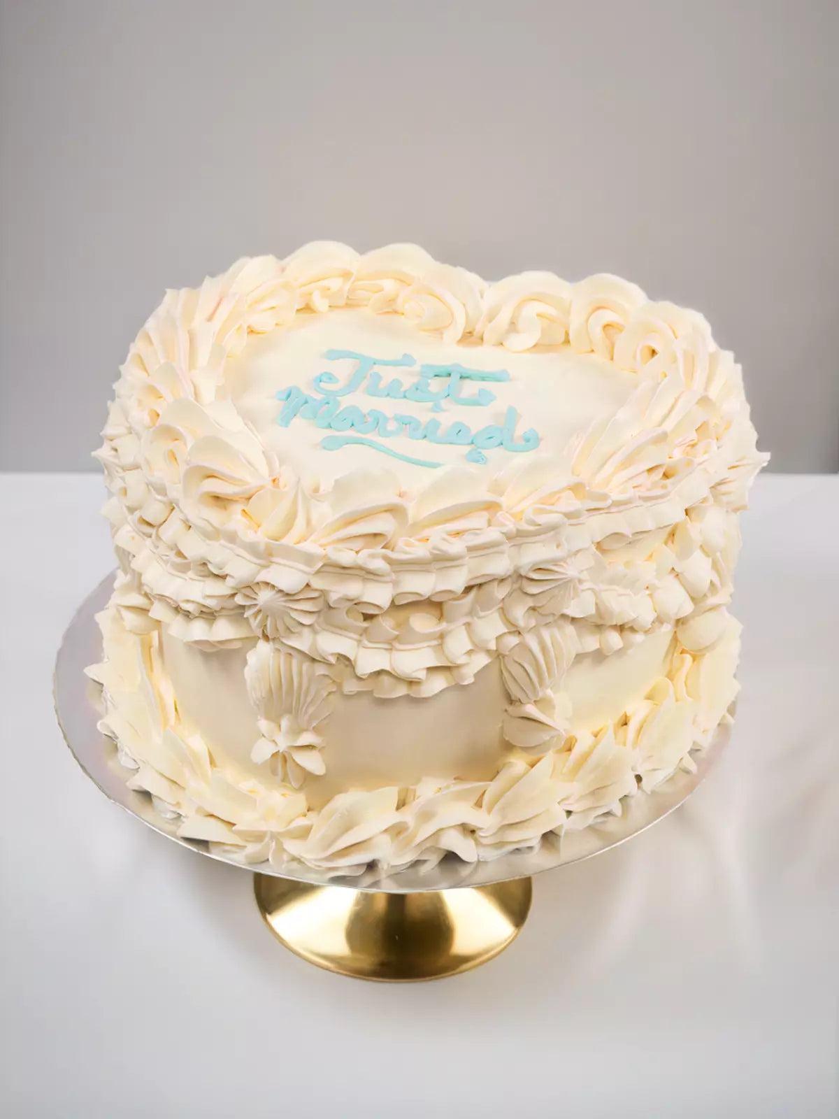 The Custom Registry Wedding Cake is heart-shaped with ornate frosting borders, seashell decorations, and light blue icing. Displayed on a gold cake stand, this personalized "Just Married" cake adds a charming touch to your celebration.