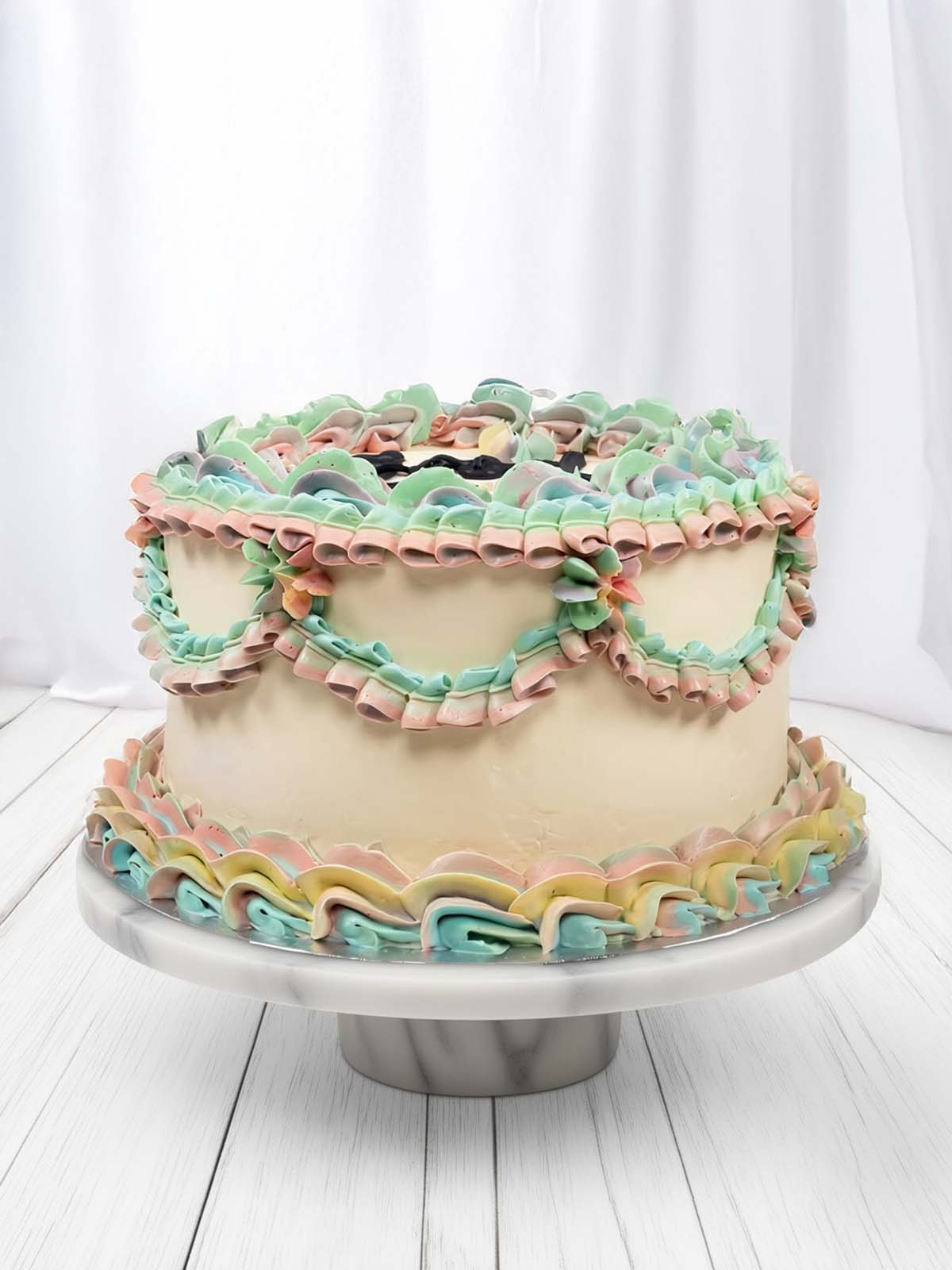 Round cake with pastel pink, blue, green, and yellow ruffles on a white marble cake stand
