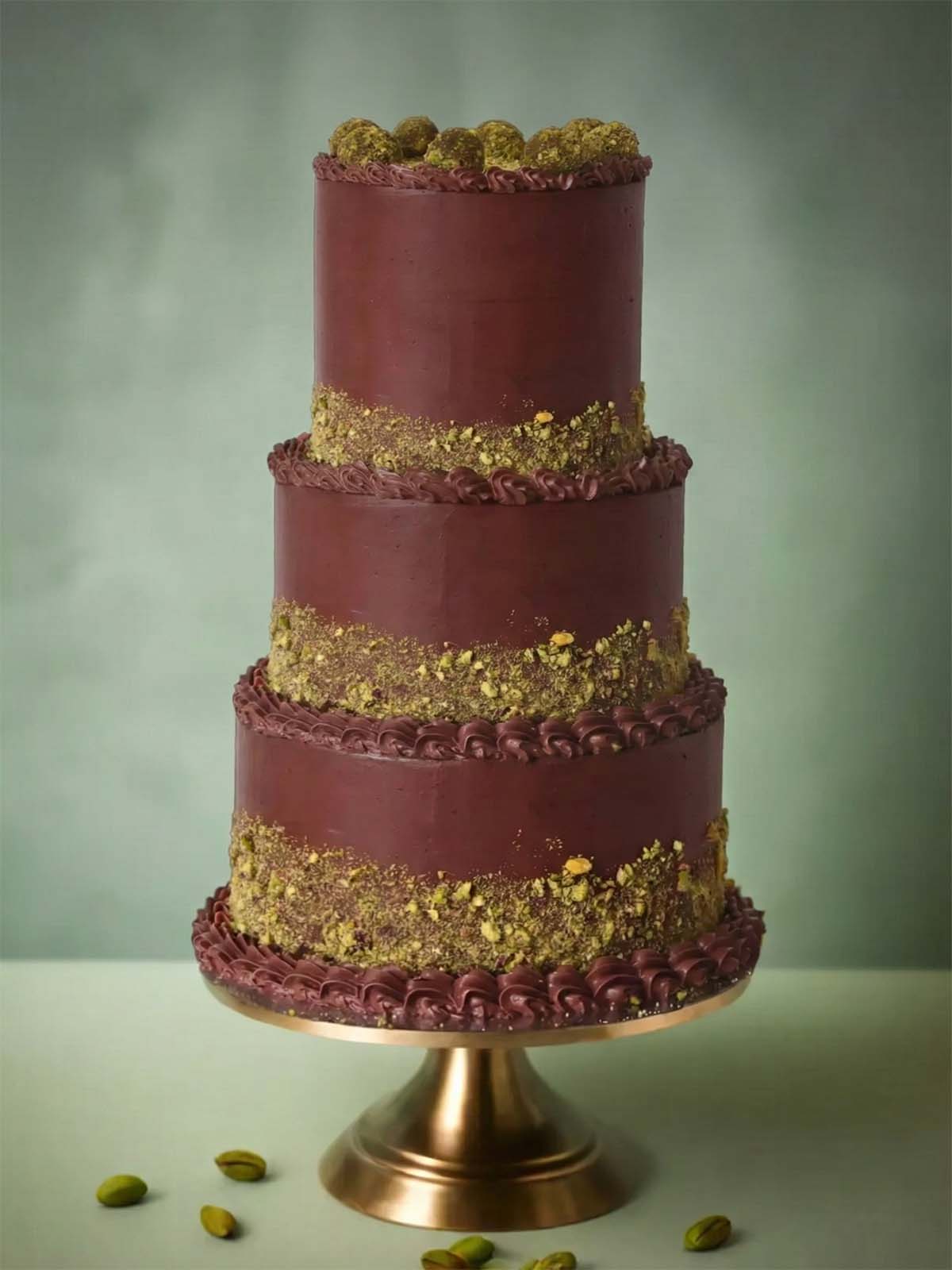 A bespoke three-tier dark chocolate wedding cake featuring textured pistachio crumb bands and decorative piping, displayed on a gold stand for a London wedding.