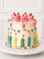 Full view of a playful birthday cake decorated with hand-piped pink pigs, party hats, pastel bunting, balloons, candles and sprinkles.