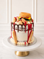 Quirky fast-food illusion cake featuring a red velvet sponge base, Belgian chocolate ganache drip, and an edible doughnut burger with a brownie patty, fondant cheese, and biscuit chips. Delivered across Surrey.