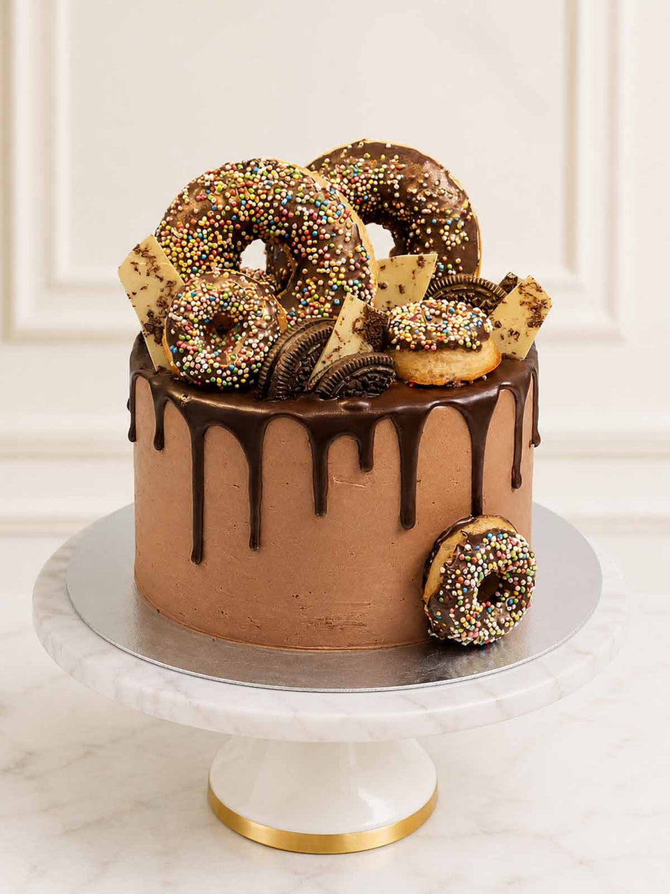 A decadent chocolate and doughnut celebration cake featuring chocolate-glazed buttermilk doughnuts, golden Oreos, and white chocolate shards dusted with gold lustre, delivered in London.