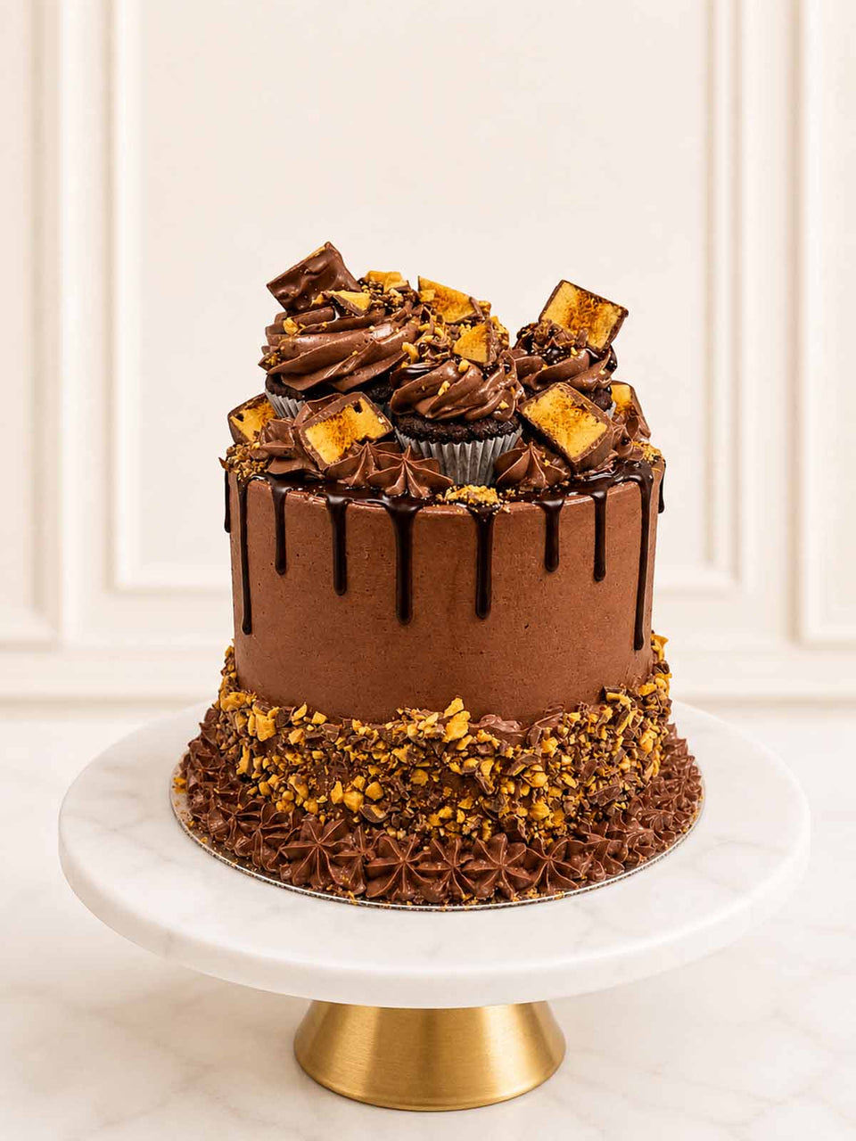 Indulgent three-layer chocolate sponge celebration cake frosted with chocolate Swiss meringue buttercream, topped with honeycomb chocolate bars, glossy Belgian chocolate drip, and mini cupcakes. Delivered in London.