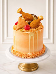 A vibrant Diwali celebration cake featuring orange and green buttercream, white chocolate raita drip, and edible cake-fudge samosas and chicken leg toppers. Perfect for Festival of Lights parties. Delivered in London.