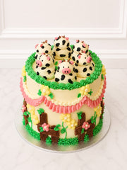 Farmyard-style Kitsch Cow Cake topped with buttercream cows and finished with piped green grass, pastel ruffles, blossoms and fencing.
