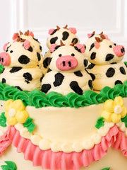 Close-up of hand-piped buttercream cow toppers on a vanilla Swiss meringue buttercream cake with green piping and pink ruffles.