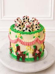 Angled view of a Kitsch Cow Cake decorated with hand-piped buttercream cows, green piping, pink ruffles, yellow blossoms and chocolate fencing.