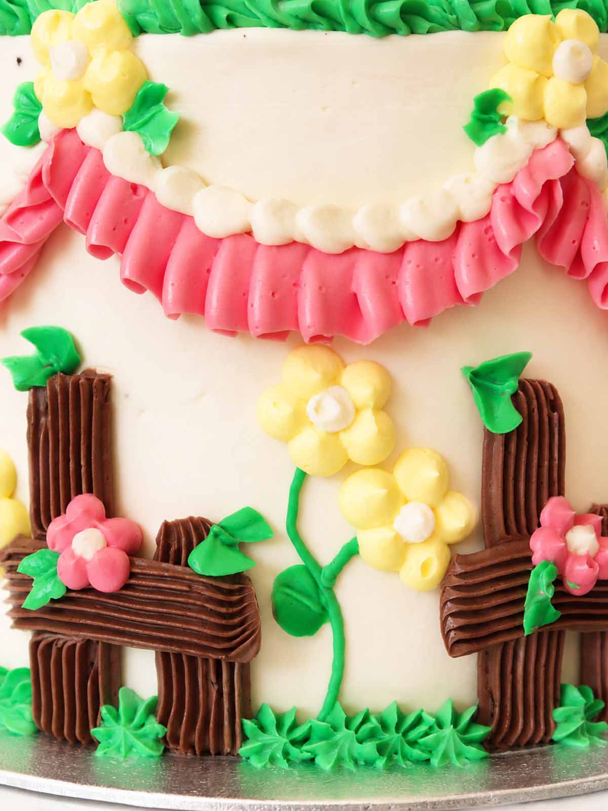 Close-up detail of the Kitsch Cow Cake showing hand-piped pink ruffles, yellow blossoms, green vines and chocolate-style buttercream fencing.
