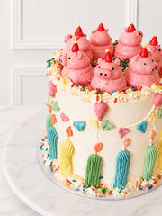 Angled view of a kitsch celebration cake topped with pink buttercream pigs and decorated with bunting, balloons, candles and sprinkles.