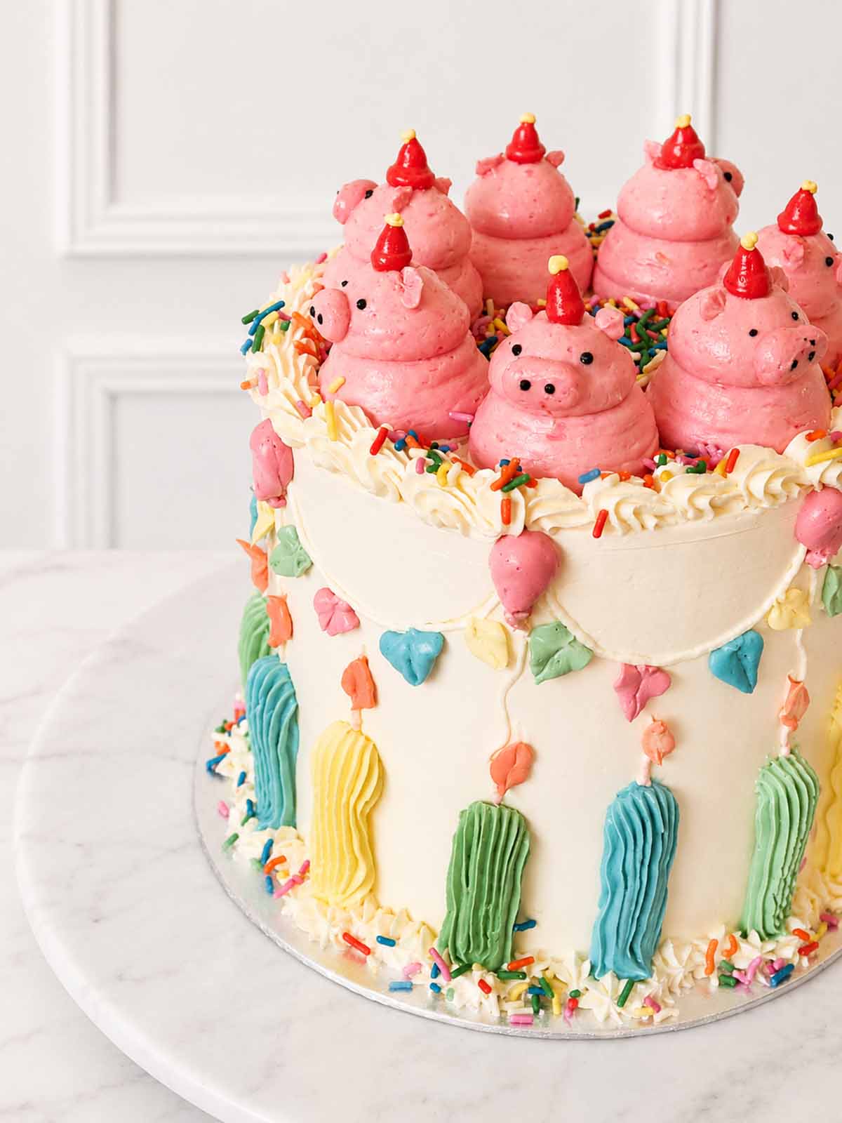 Angled view of a kitsch celebration cake topped with pink buttercream pigs and decorated with bunting, balloons, candles and sprinkles.