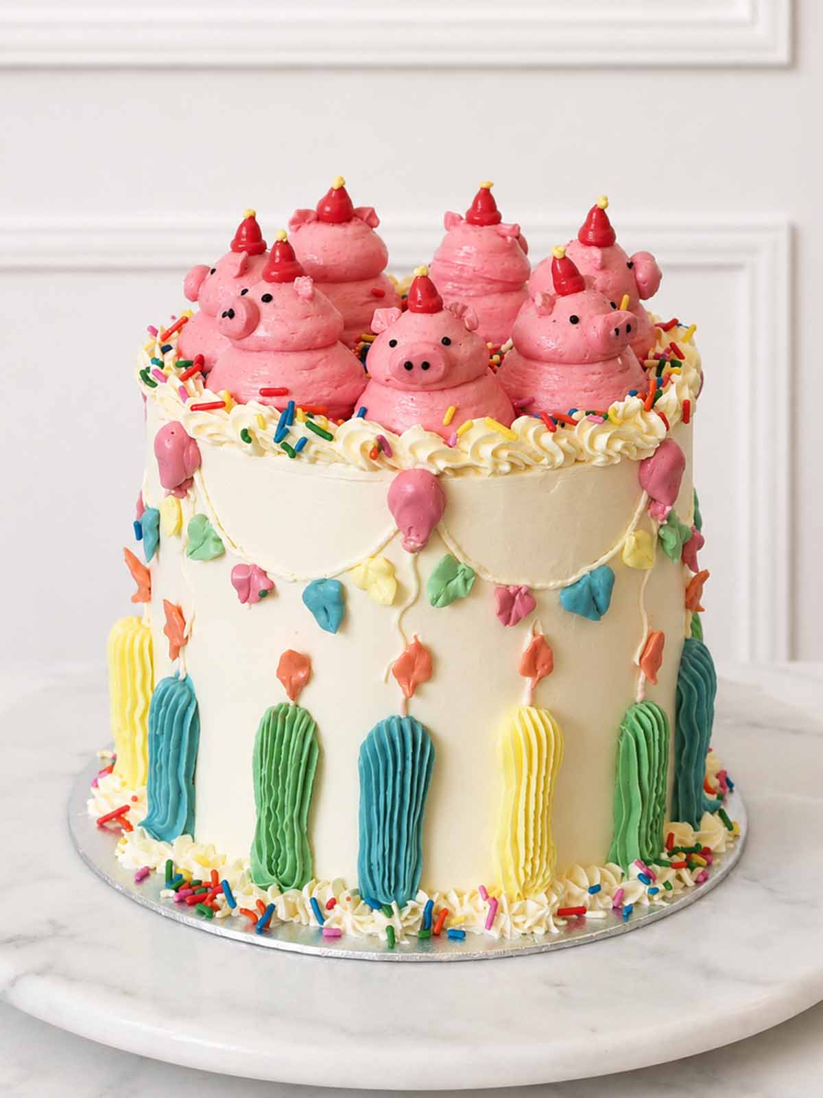 Front view of a tall vanilla buttercream cake with pink party pigs, colourful bunting, balloons, candles and rainbow sprinkles.
