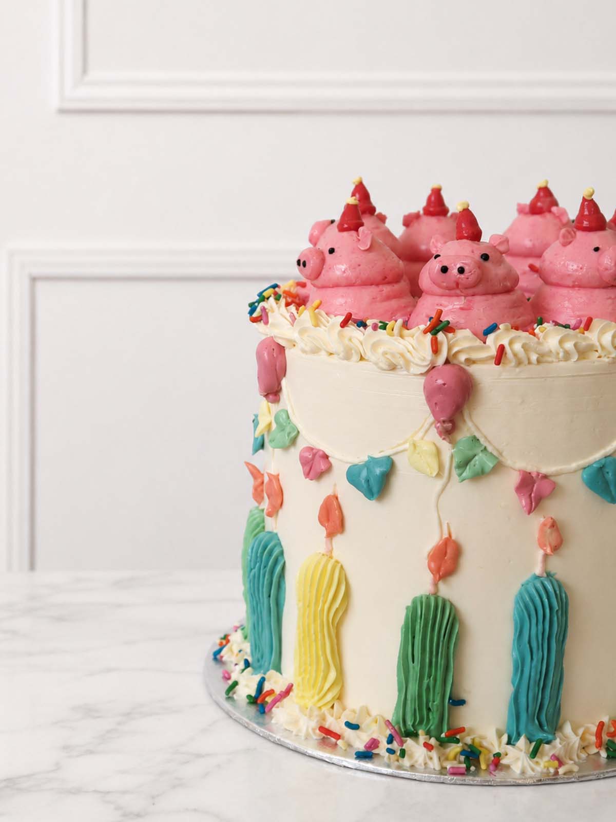 Kitsch celebration cake decorated with hand-piped pink buttercream pigs, party hats, bunting, balloons, candles and rainbow sprinkles.