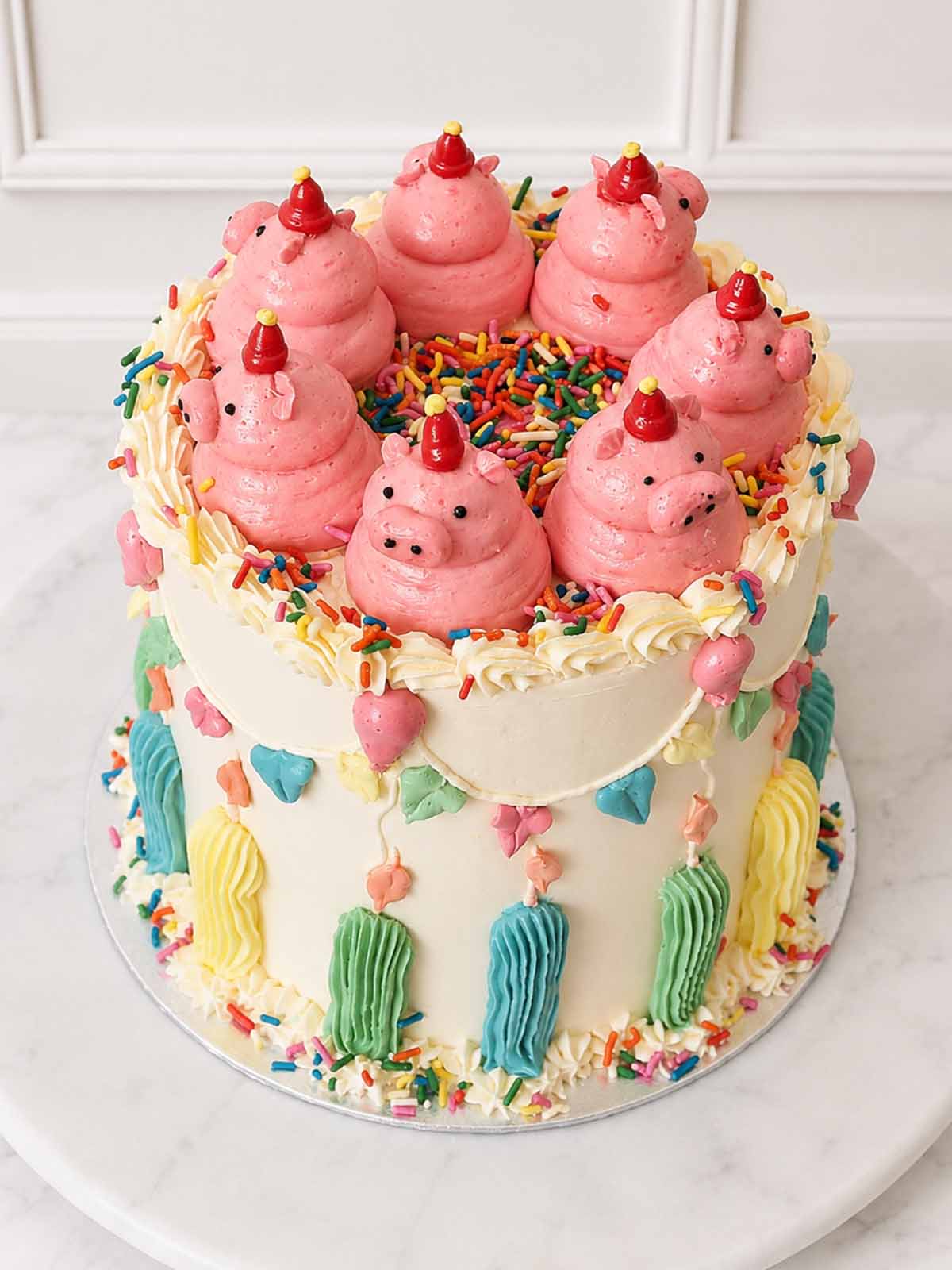 Elevated view of a vanilla celebration cake topped with hand-piped buttercream pigs in party hats and a centre filled with sprinkles.