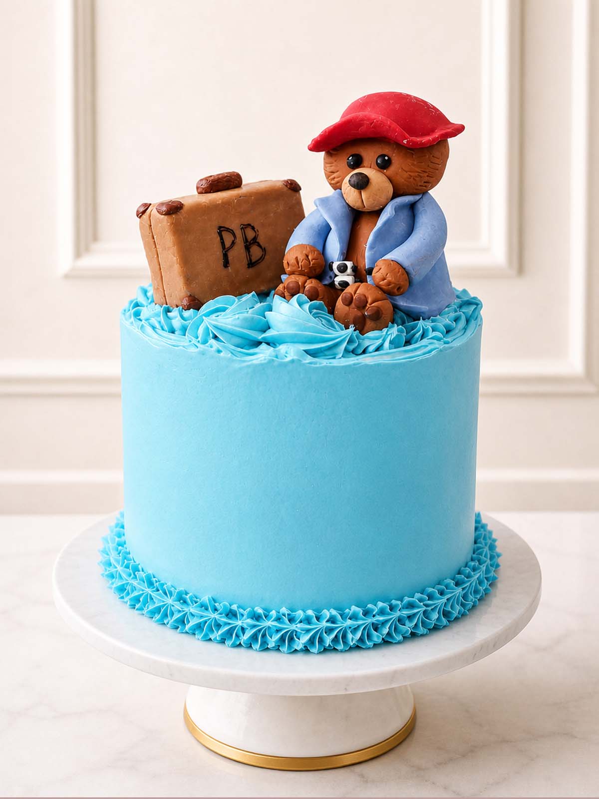 Whimsical storybook-inspired birthday cake featuring a hand-sculpted sugar paste bear with a raggedy suitcase, smooth blue Swiss meringue buttercream, and three layers of vanilla sponge. Perfect for toddler parties and London-themed celebrations.