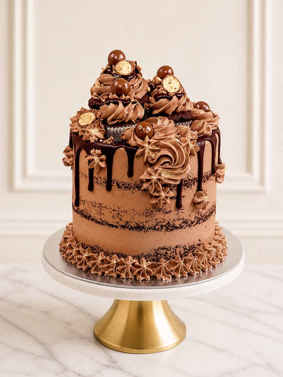 A fun, malted milkshake-inspired celebration cake featuring three layers of chocolate sponge, semi-naked malt-chocolate Swiss meringue buttercream, and a glossy Belgian chocolate drip. Topped with crushed Maltesers and milkshake-style cupcakes. Delivered in London and Surrey.