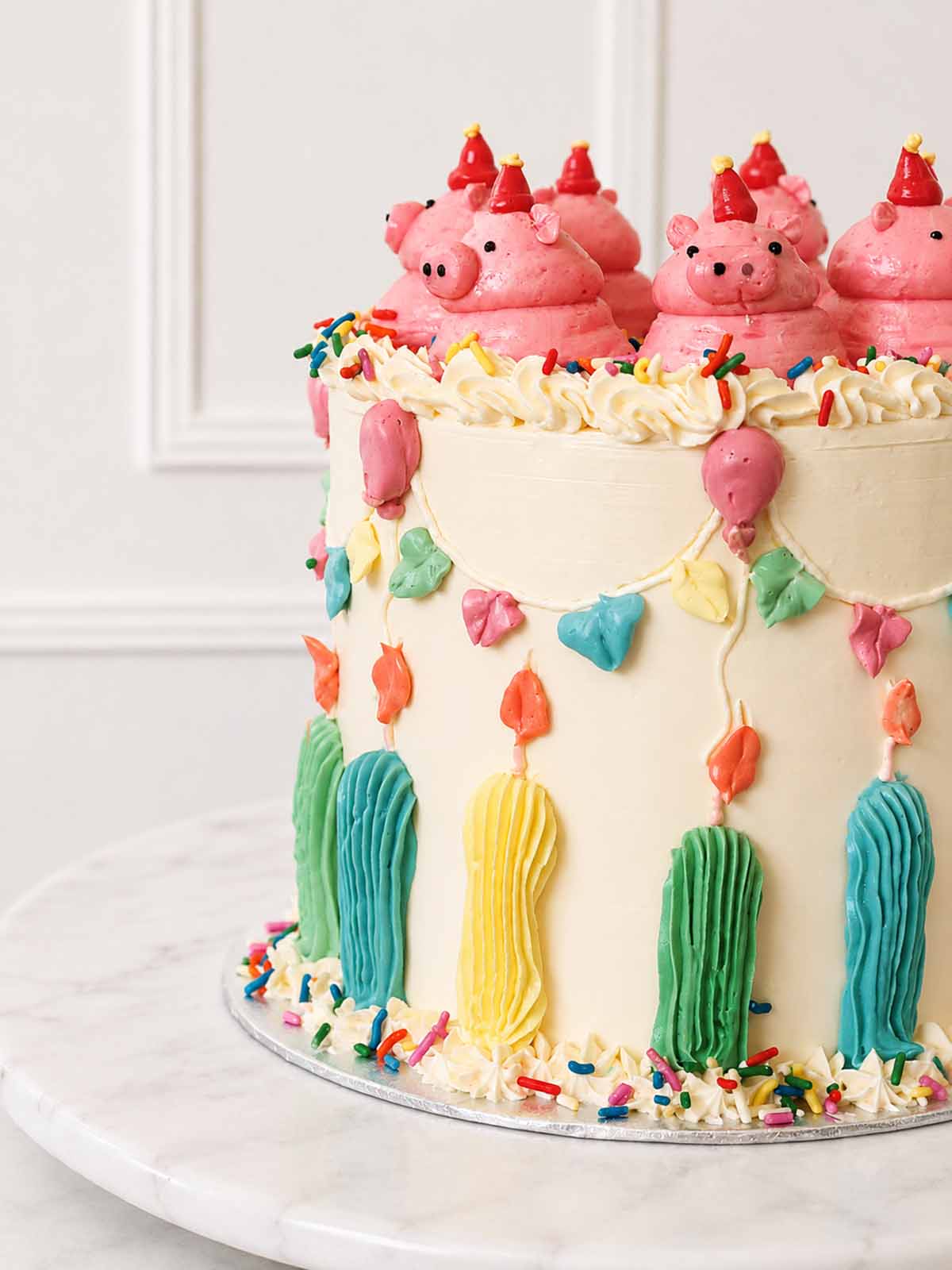 Close-up of a kitsch pig party cake with pink buttercream pigs, red party hats, piped bunting, balloons and colourful candles.