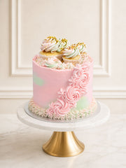 A romantic rose and pistachio celebration cake featuring pink rose-infused Swiss meringue buttercream, pistachio sponge, and a crown of mini cupcakes and fresh raspberries, delivered in London.