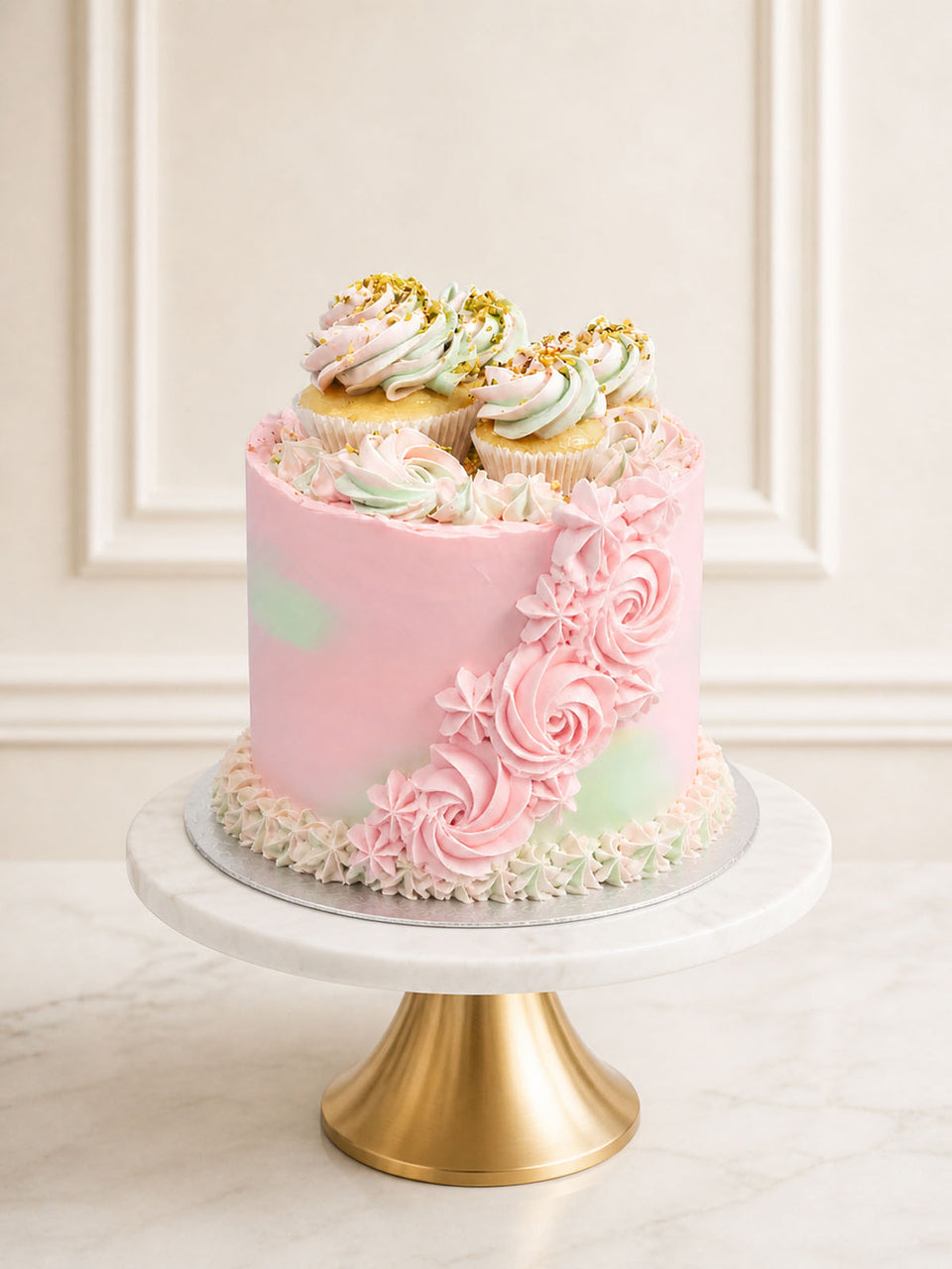 A romantic rose and pistachio celebration cake featuring pink rose-infused Swiss meringue buttercream, pistachio sponge, and a crown of mini cupcakes and fresh raspberries, delivered in London.