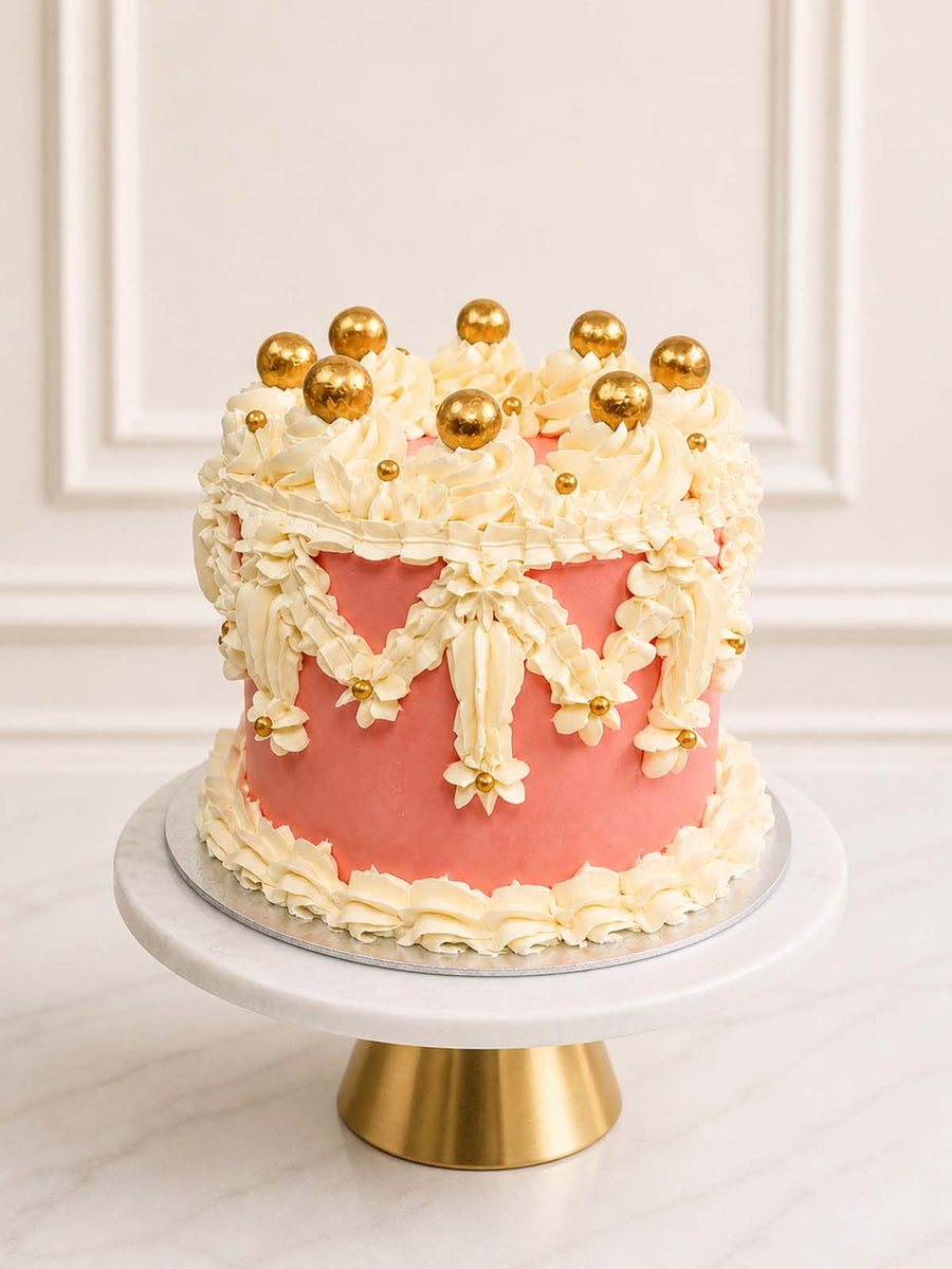 A majestic pink and ivory celebration cake inspired by Windsor Castle featuring three layers of vanilla sponge, intricate vintage buttercream piping, and golden chocolate pearls