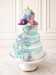 Whimsical romantic wedding cake featuring a serene blue watercolour buttercream finish, beautifully decorated with a gold-gilded birdcage, hand-modelled fondant love-birds, and delicate hydrangeas. Delivered across Surrey.