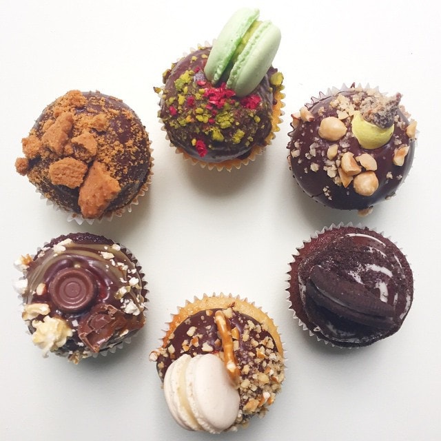 Six cupcakes with various toppings including macarons, nuts, cookies, and chocolate