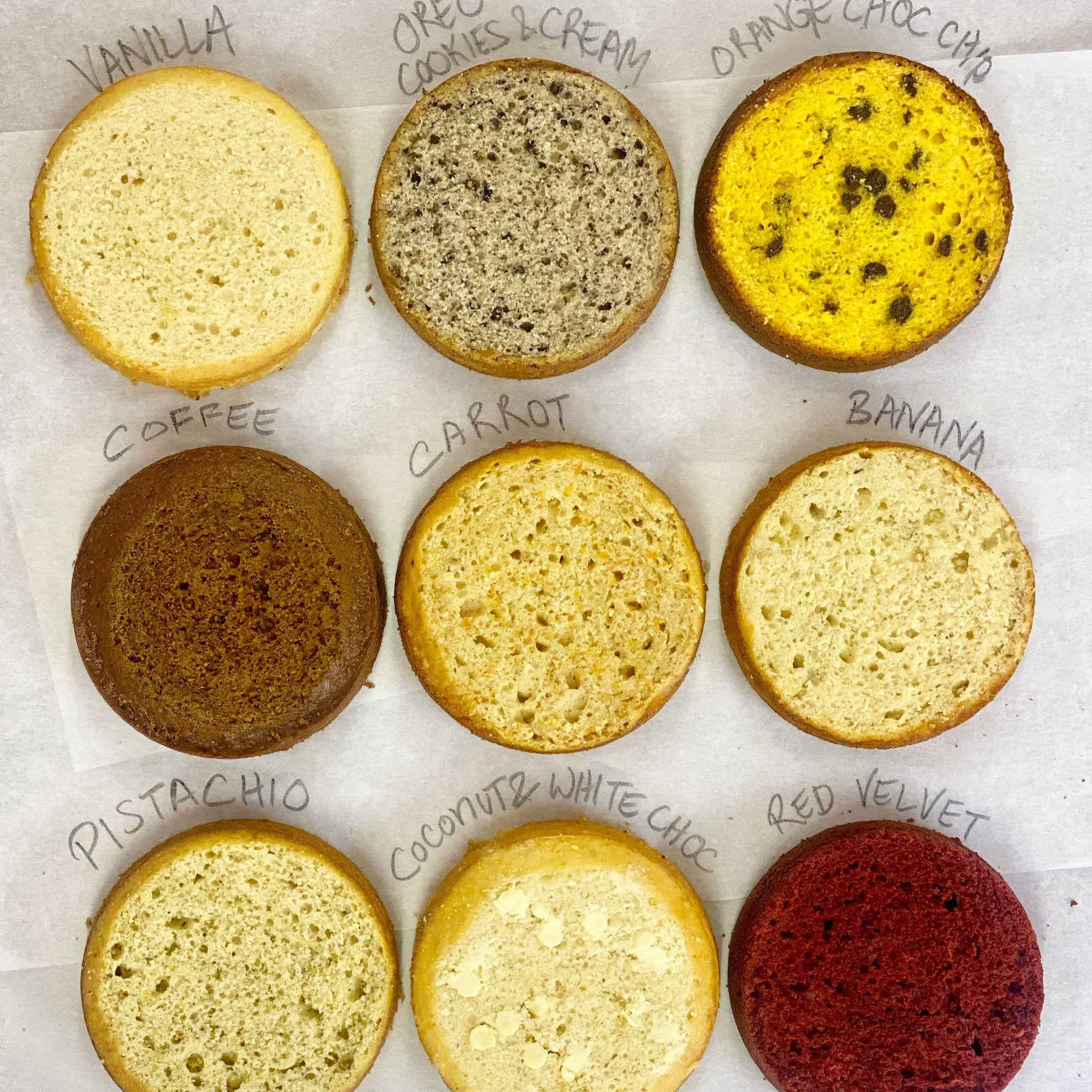Nine round cake layers, each a different flavor from the Original Hero Sponge Recipe, are arranged in a 3x3 grid on parchment: vanilla, cookies & cream, orange choc chip, coffee, carrot, banana, pistachio, coconut white choc, and red velvet.